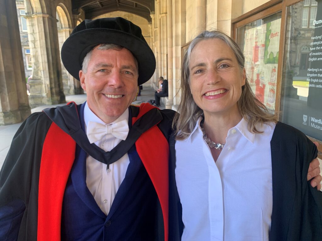 A male University professor in gown and hood, with a female graduate in gown and white shirt