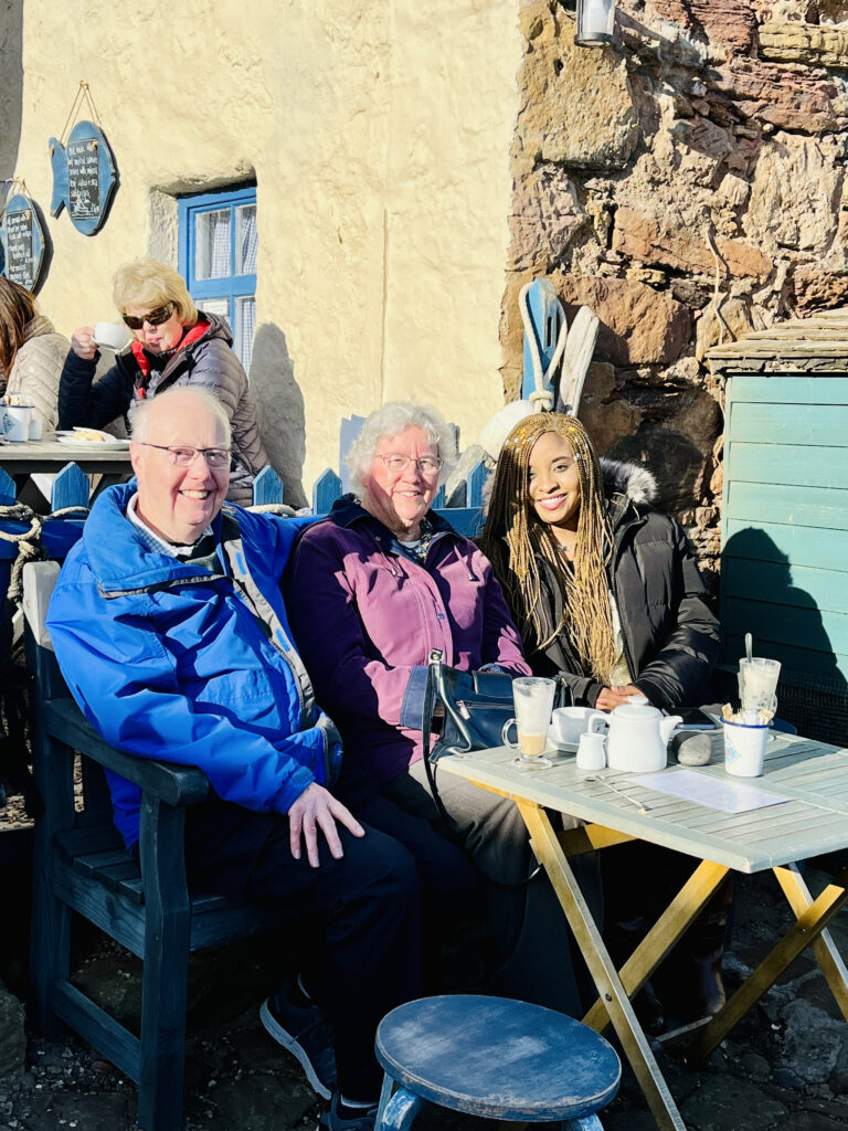 The trio sit outside in a local cafe - a table full of tea and coffee sits in front of them. The three sit side by side on a bench, smiling in the sunshine