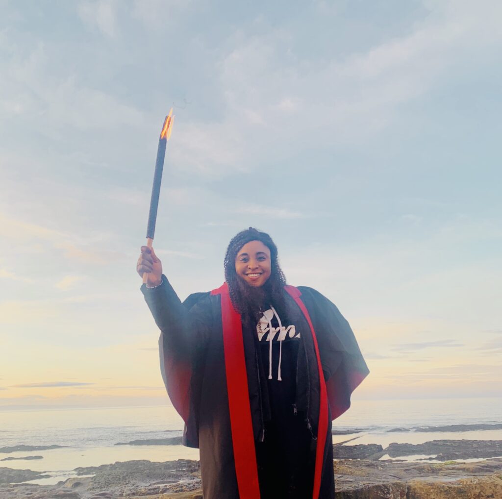 Racheal stands smiling in her postgraduate gown, holding a lit torch. In the background we can see the sea at sunset