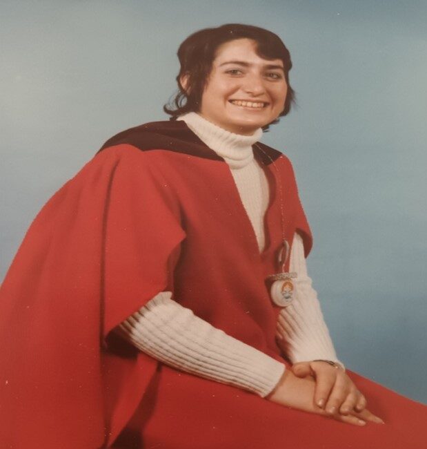 Jenny poses for an official University portrait in her red gown in front of a pale blue background.