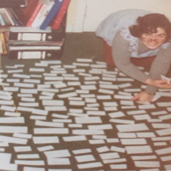 Jenny looks up at the camera smiling, she is on the floor sorting lots of small pieces of paper.