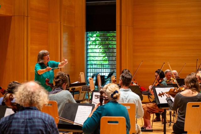 Jill conducts an orchestra in the McPherson Recital Room in the Laidlaw Music Centre. The musicians are visible but mainly with their backs to the camera - Jill is position to the left, waving her arms to conduct. 
