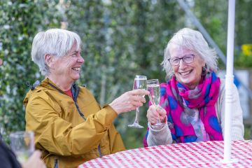 Anne and Jenny sit at a table outdoors, they are both smiling and clinking glasses of prosecco with one another.