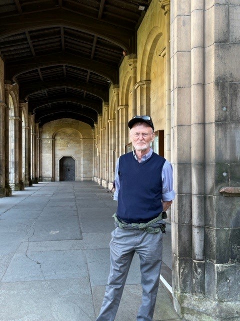 The image shows a white, older male with white hair and cap, grey trousers and blue knitted tank top with blue shirt, standing in a historic grey-stone cloister.