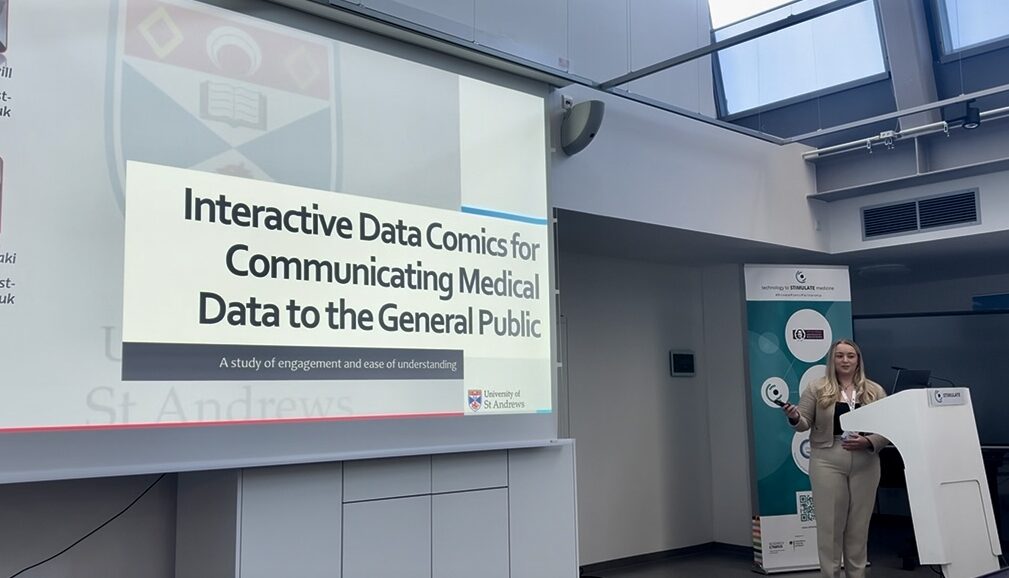Melissa is presenting from a stand in the bottom right of the picture. To the left we can see a projector screen with a slide reading 'Interactive Data Comics for Communicating Medical Data to the General Public, A study of engagement and ease of understanding'.
