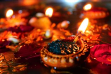 The image is red, orange and black in colour and shows a photograph of a diya, a small oil lamp used to celebrate Diwali. The lamp is alight.