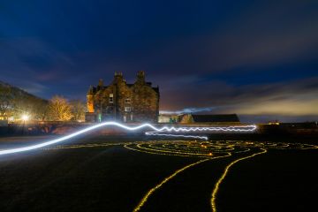 The image is taken in the dark of night and shows a light trail through the sky above a maze made of tealights.