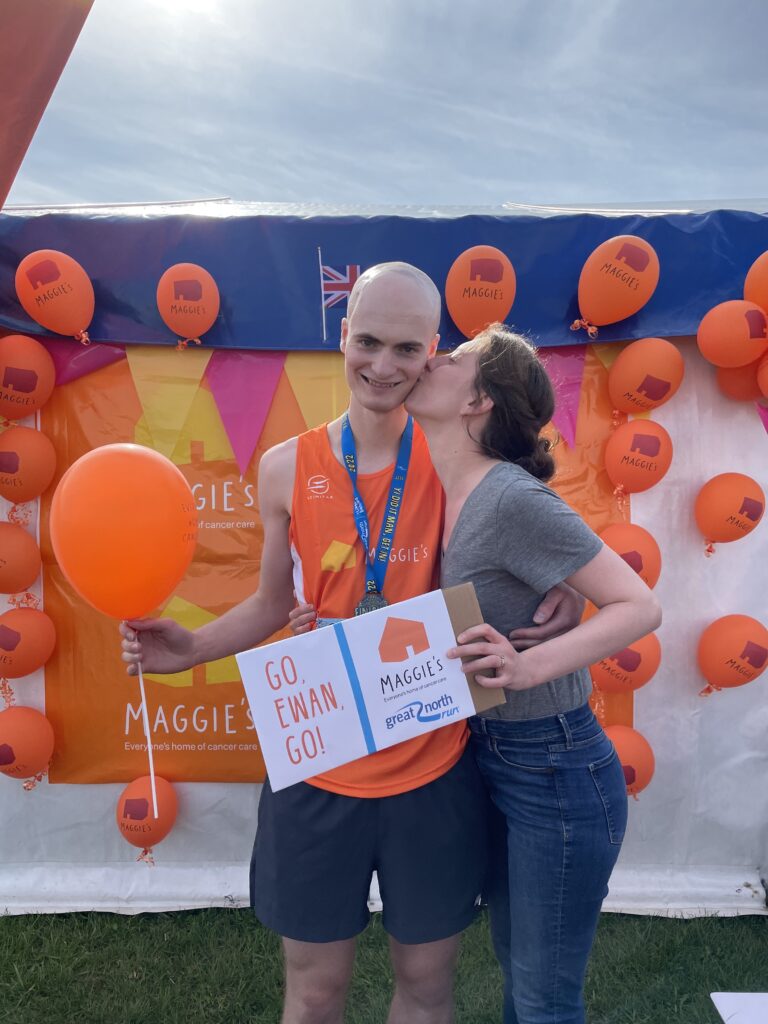 Ewan and Karlee at the half-marathon. Ewan is smiling at the camera, wearing running kit and a medal. Karlee is kissing Ewan on the cheek while holding a sign that says 'GO EWAN GO!' with a Maggie's banner and balloons in the background.