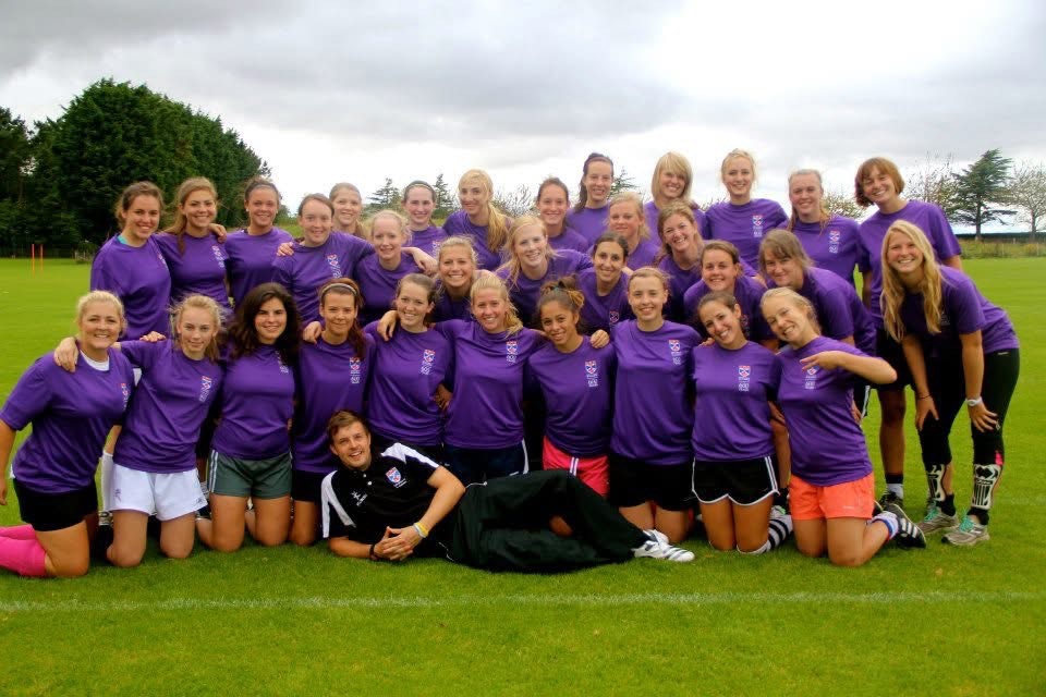 The Women's Football Club pose wearing matching purple sports shirts on a grassy field, with the coach lying in the front.