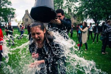 Collette being drenched with water from a black bucket, surrounded by people on a grassy area with trees. This is part of the traditional 'soaking' at St Andrews.
