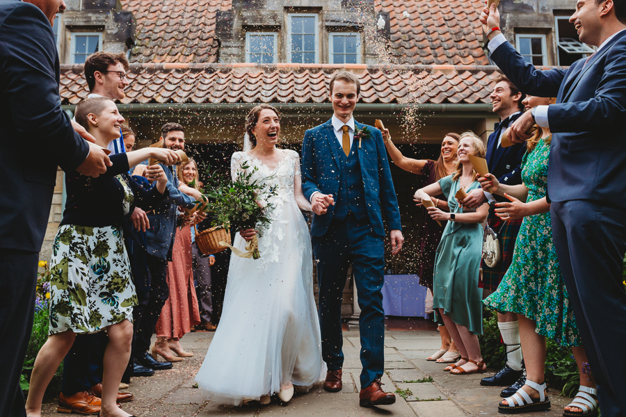 Karlee and Ewan walk through confetti-throwing guests in the courtyard of All Saints' Church. Karlee wears a white wedding dress and holds a bouquet of flowers, Ewan wears a blue suit with a yellow tie - the pair are holding hands and smiling. 