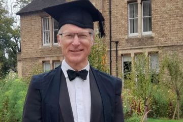 A man wearing glasses, a black bow tie, black suit jacket, white shirt and mortar board stands smiling towards the camera. Behind him is a red brick house and green leafy hedges.