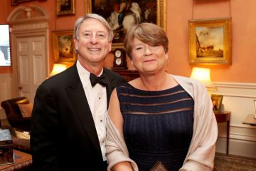 A man and a woman in formal Black Tie attire, pictured in a warmly lit historic drawing room.