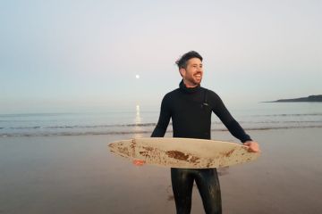 Dr Chris McKnight wears a black wetsuit holding a white surfboard on a sandy beach at sunset, with calm waves, the moon reflecting on the water.