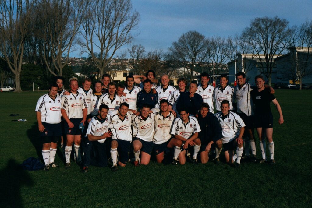 The St Andrews Men's Rugby Team pose for a photo on the pitch. 18 team members pose in the back, with 7 kneeling in the front row.