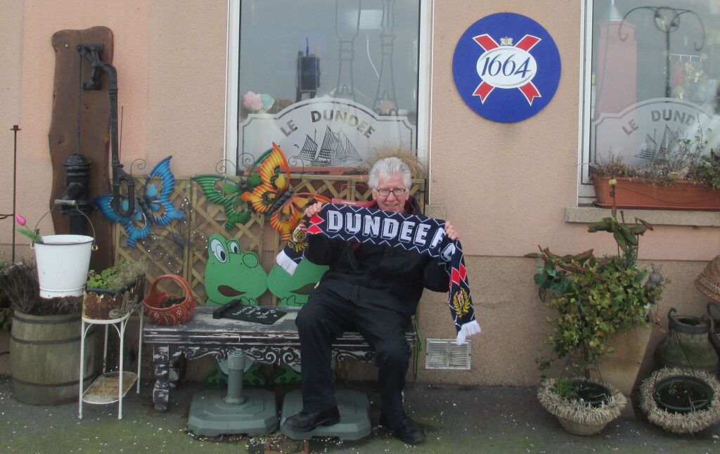 JP Creton sits on a bench outside a bar in a French town, holding up a Dundee Football Club scarf.