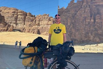 Connor McElwaine, standing in a yellow t-shirt, sunglasses and cycling gear, with his bicyle, outside Maraya, which is the world's largest mirrored building.