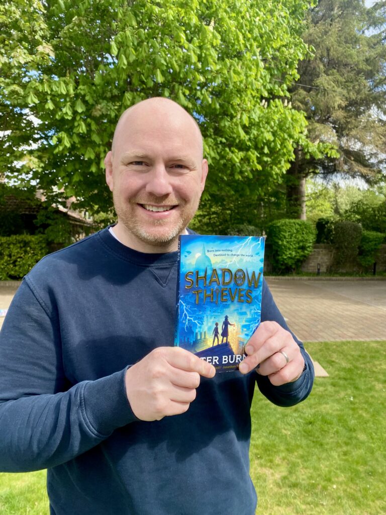 Paul stands outside with a background of trees and foliage, smiling holding his book, Shadow Thieves, up to the camera.