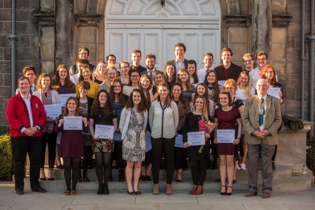 Participants in the 2018 Gives Back Awards pose smiling for a group photo holding their award certificates on the steps in St Salvator's Quad. 