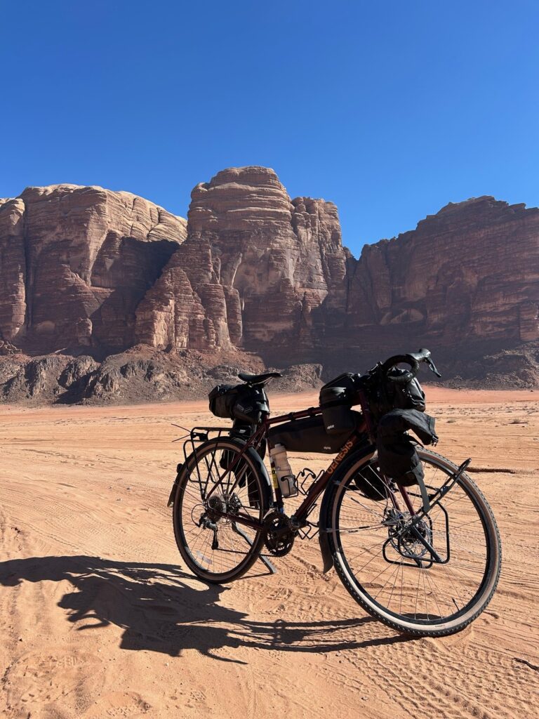 A bicycle with bags propped up in the foreground, with sand and carved rock of a desert landscape surrounding it.