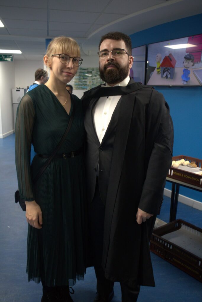 Andrei and his wife pose for a photo on graduation day, Andrei is wearing a suit with his graduation gown. 