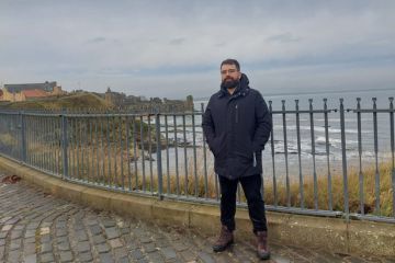 Andrei poses for a photo on the shores of St Andrews - the sea and castle can be seen in the background