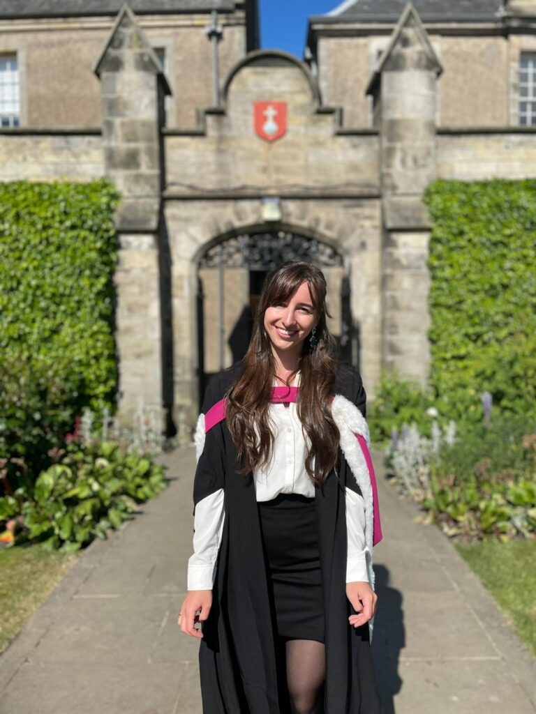 Patricia stands smiling in her graduation gown and academic dress, she is in St Salvator's Quad on a sunny day