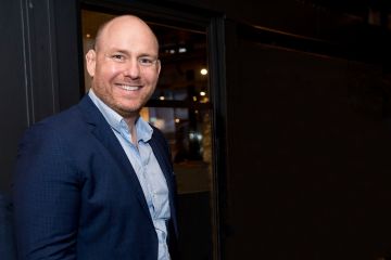 Peter stands smiling for a photo, he is dressed in a navy blue suit with a blue shirt