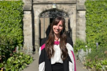 Patricia stands smiling in her graduation gown and academic dress, she is in St Salvator's Quad on a sunny day
