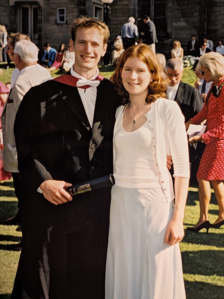Ed and Helen pose for a photo in a bustling St Salvator's Quad. Ed is wearing graduation robes and holding his degree scroll