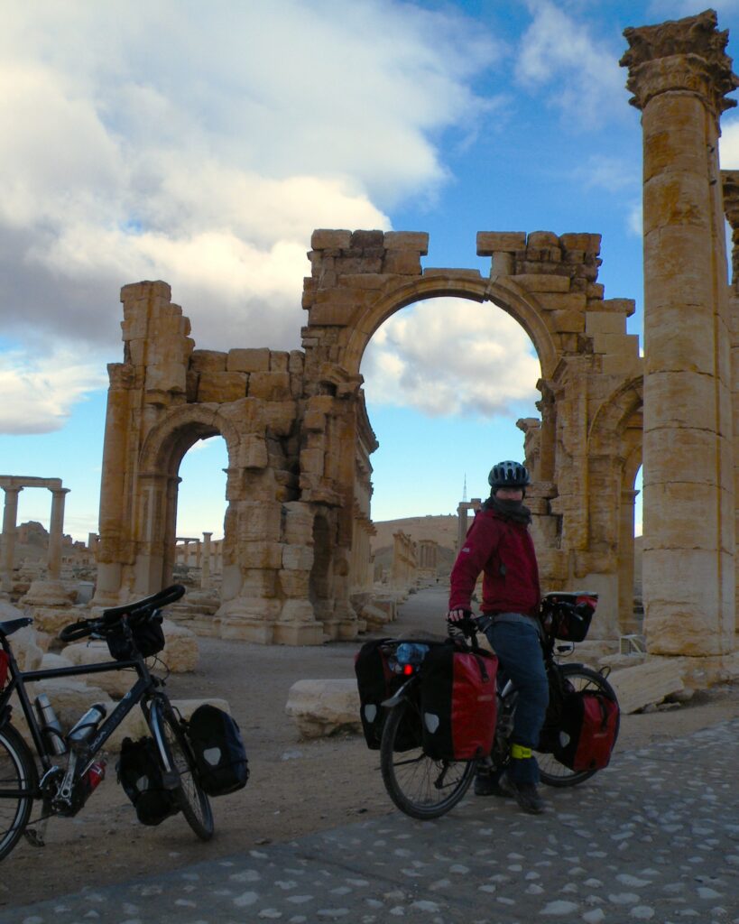 Helen sits on her bike, with Ed's bike propped up to her left. The Monumental Arch of Palmyra, Syria can be seen in the background. 