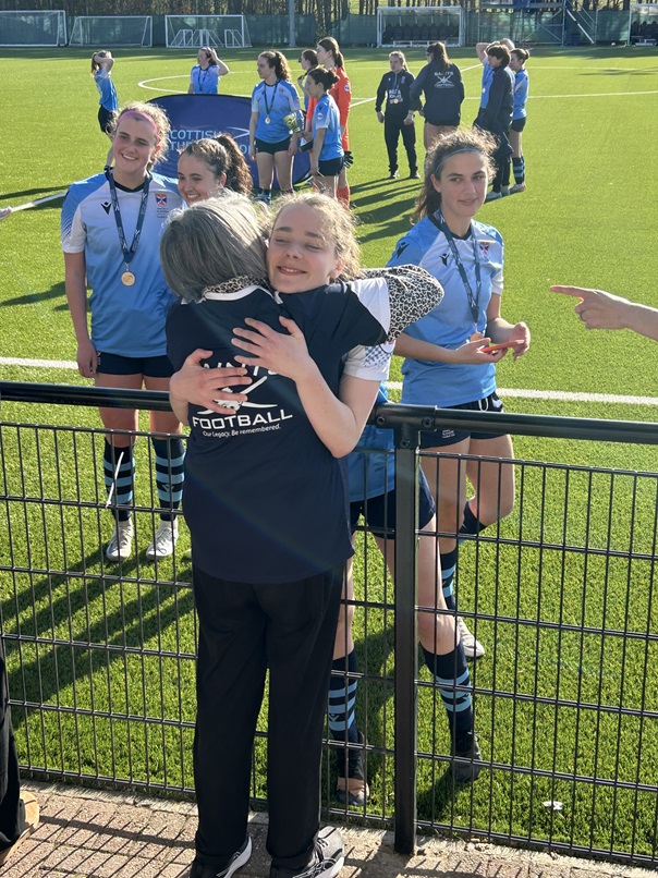 A grandmother, Fiona Goodchild, hugs her granddaughter as they celebrate a win for her football team. The young female footballers are in a blue strip on the pitch.
