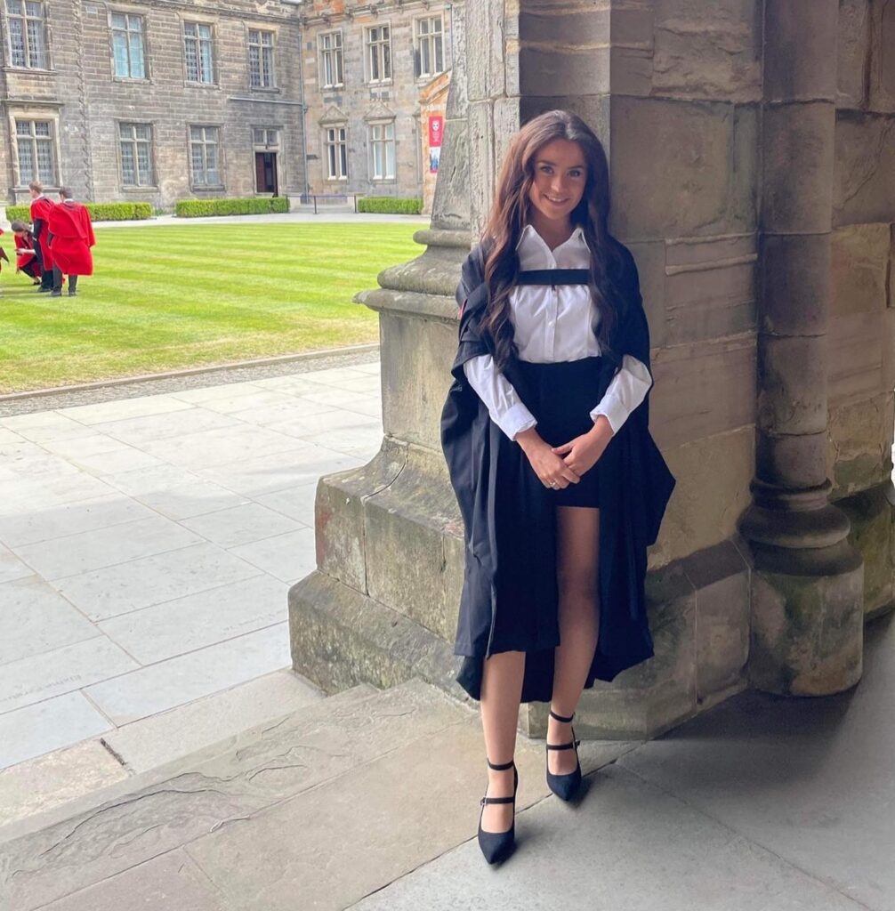 Laura poses for a photo in her graduation gown and academic dress. She stand in the cloisters, with a view of St Salvator's Quad in the background. 