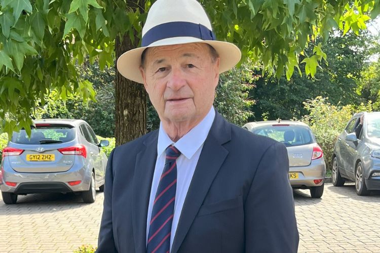 A gentleman smartly dressed in a suit, tie and panama hat stands in front of a tree in the sunshine