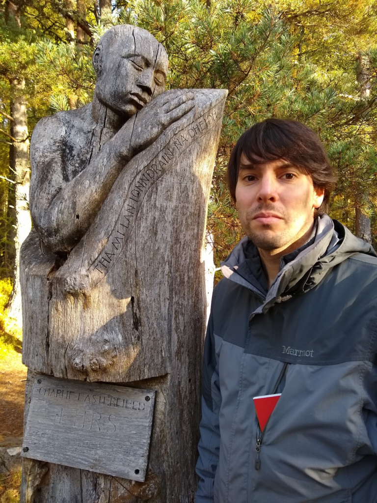 Dr Paul Flaig looks directly at the camera, a wood statue is to his left. In the background we can see that Paul is in a forest.
