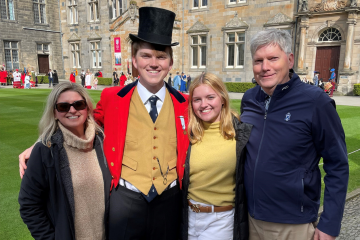Four people, Edward, Izzie, Carrie, and Lyon, stand smiling for a photo in St Salvator's Quad. Lyon is wearing historical dress and a top hat as his character 'Sir Hugh Lyon Playfair'
