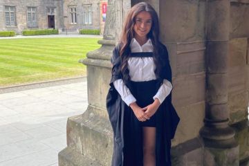 Laura poses for a photo in her graduation gown and academic dress. She stand in the cloisters, with a view of St Salvator's Quad in the background.