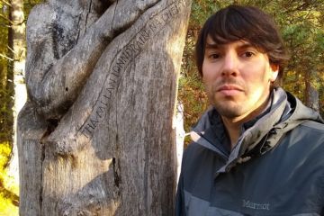 Dr Paul Flaig looks directly at the camera, a wood statue is to his left. In the background we can see that Paul is in a forest.