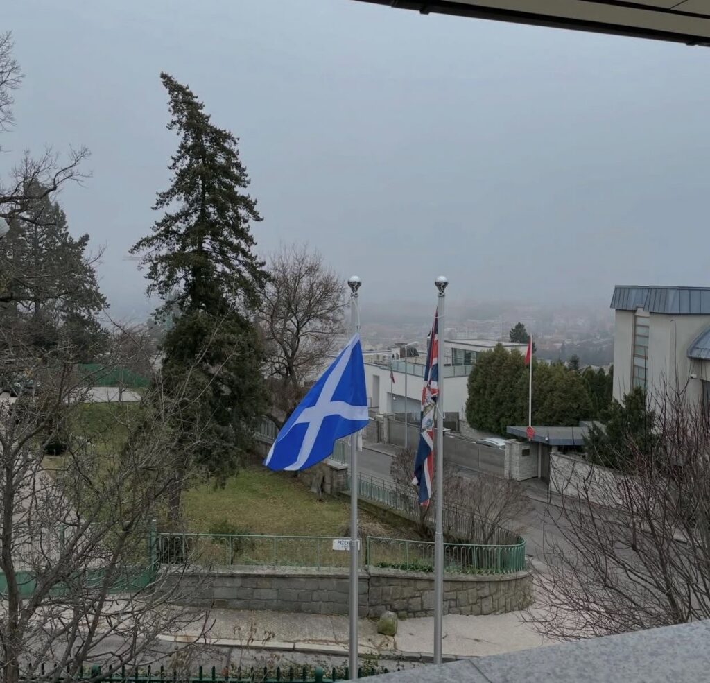 A Scottish and British flag fly on flagpoles, the landscape of Slovakia is obscured by fog