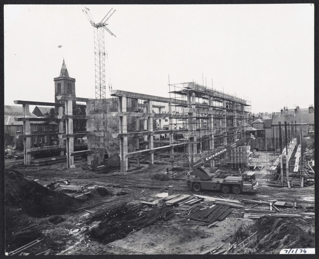 The Main Library building under construction, surrounded by scaffolding, a tower crane and a mobile crane. Building materials cover the muddy site, with St Salvator's visible in the background
