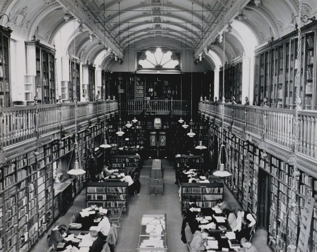 A black and white overhead shot of the Arts and Divinity Reading Room. The image shows floor-to-ceiling bookshelves, ornate arched ceilings, hanging lamps and readers seated at long tables.