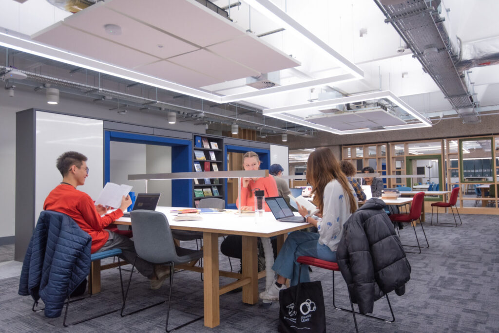 Students study around a large shared table in the Main Library, reading papers and using laptops beneath rectangular ceiling lights. Bookshelves and additional study areas are visible in the background.
