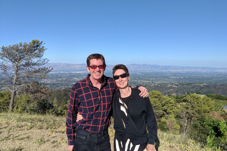 A couple, Lindsay and Nicky Grant, stand atop a hill in Silicon Valley, blue skies and vineyards behind them.