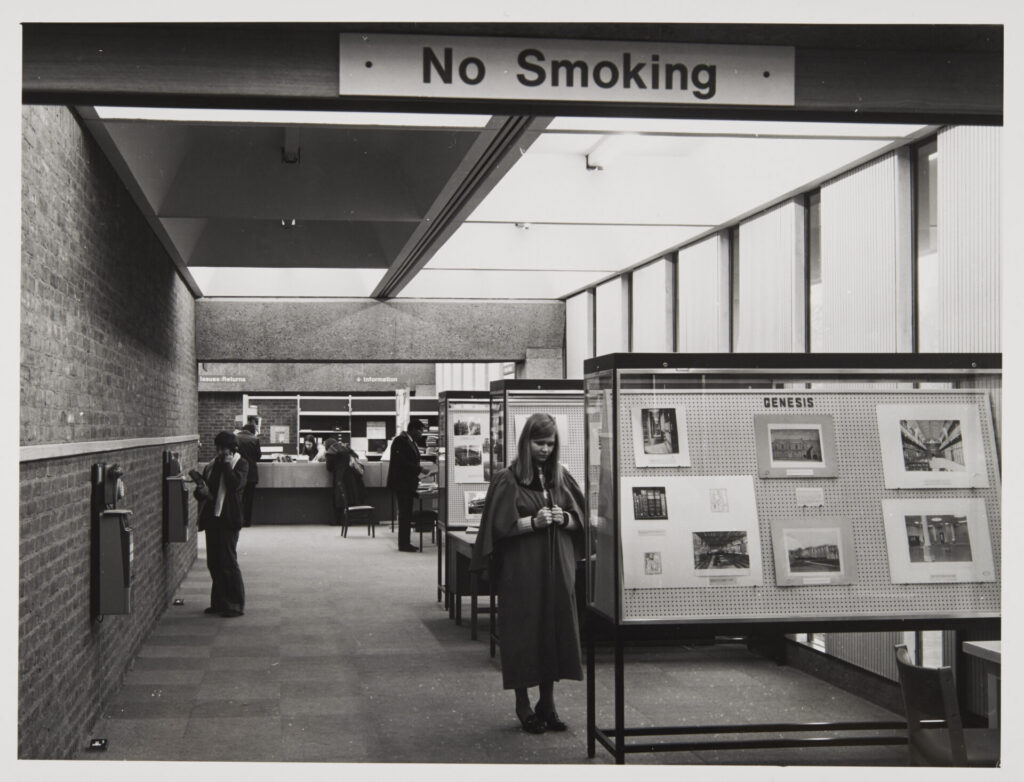 Black and white image of a student in a St Andrews gown stood in the Main Library's exhibition space. Above her head a sign reads 'no smoking'. The information helpdesk can be seen at the back of the image