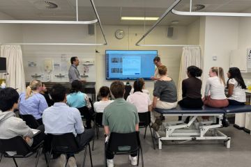 A group of St Andrews medical students in a clinical setting gather around a screen displaying the SimPatient AI