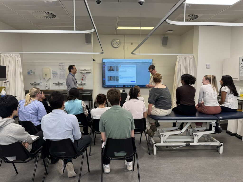A group of St Andrews medical students in a clinical setting gather around a screen displaying the SimPatient AI 