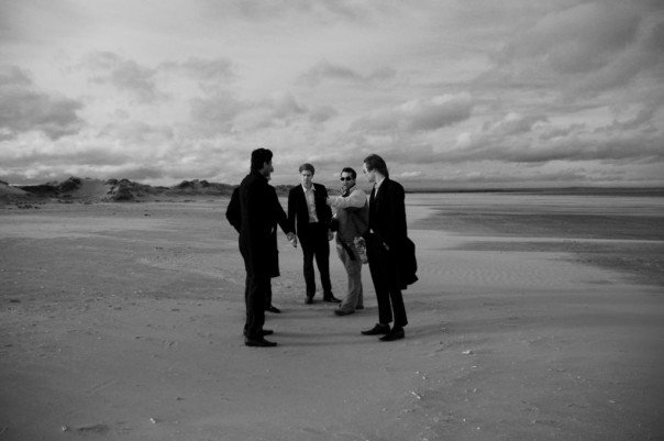 Black and white photograph, five men dressed in formalwear are stood in a semi-circle deep in conversation on West Sands