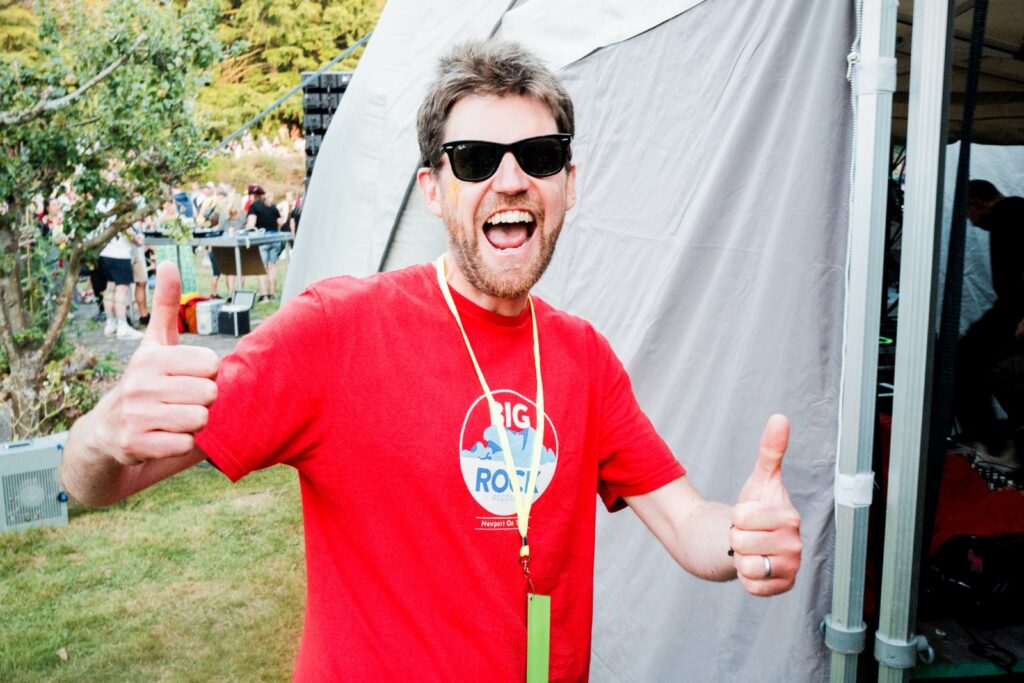 Fergus poses backstage at a music festival with both thumbs up the camera. He wears sunglasses, a lanyard and a Big Rock Records t-shirt.
