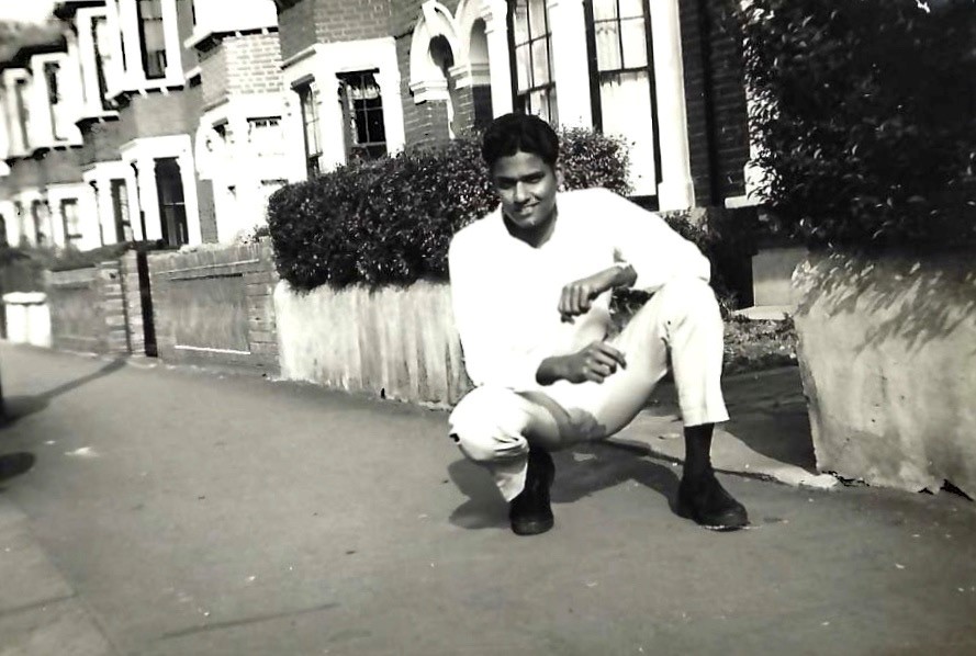 A black and white photograph of a young man from Trinidad and Tobago, crouching on the pavement next to the entrance to a terraced house.