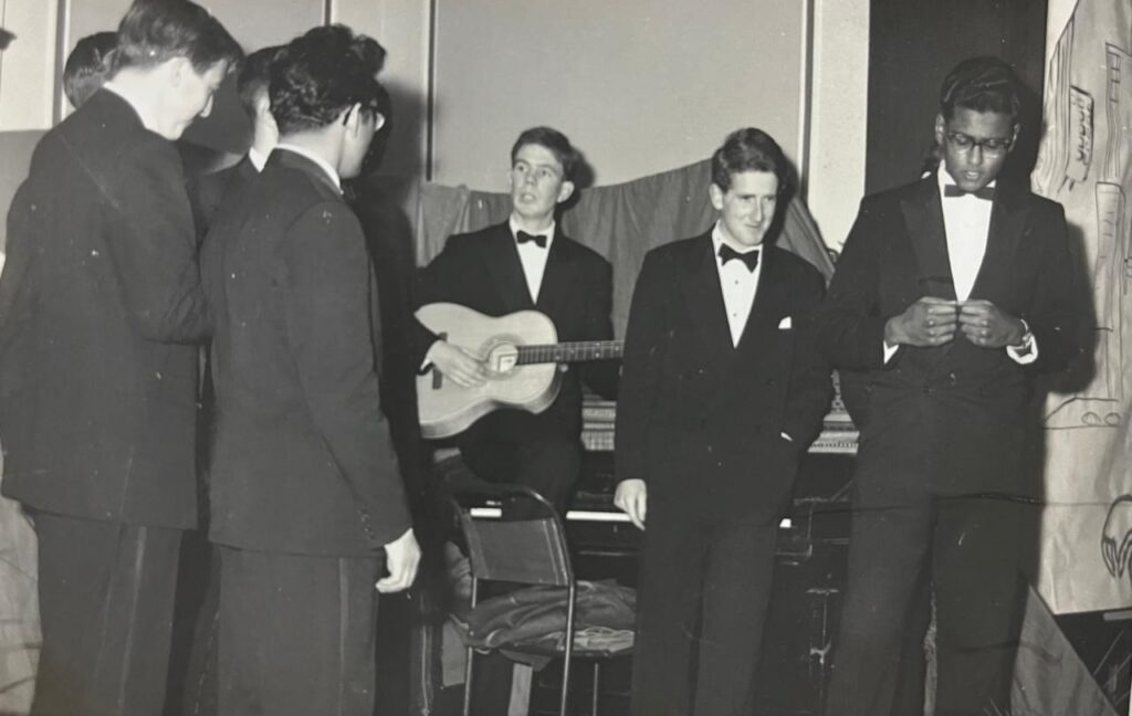 A black and white photograph from the 1960s showing a group of male students in black tie giving a musical performance. The centre student is playing a guitar.